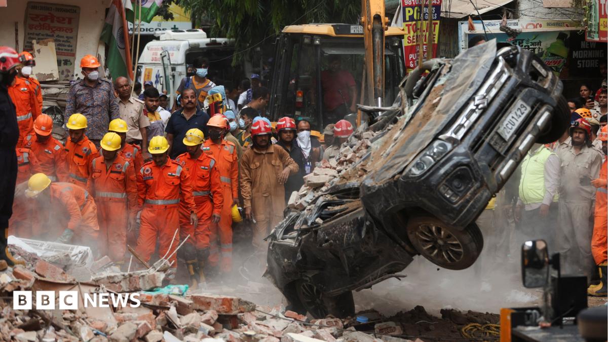 National Disaster Response Force (NDRF) personnel look on at a building collapse in Delhi, while a crushed car is pulled by a digger from the rubble.