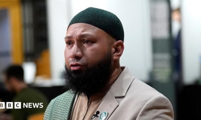 Mothin Ali looking to his right, wearing a beige suit jacket with a green and white scarf draped over his right shoulder
