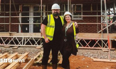 Charles and Georgina pose on a muddy building site in front of a partially completed brick house surrounded by scaffolding. Both wear safety helmets, high-visibility vests and sturdy boots. Stacks of timber lie in the foreground, while the unfinished structure, exposed windows and metal scaffold framework fill the background, illustrating the progress of the construction project.