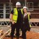 Charles and Georgina pose on a muddy building site in front of a partially completed brick house surrounded by scaffolding. Both wear safety helmets, high-visibility vests and sturdy boots. Stacks of timber lie in the foreground, while the unfinished structure, exposed windows and metal scaffold framework fill the background, illustrating the progress of the construction project.