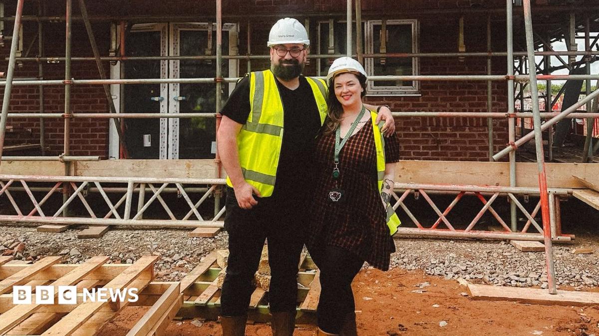 Charles and Georgina pose on a muddy building site in front of a partially completed brick house surrounded by scaffolding. Both wear safety helmets, high-visibility vests and sturdy boots. Stacks of timber lie in the foreground, while the unfinished structure, exposed windows and metal scaffold framework fill the background, illustrating the progress of the construction project.
