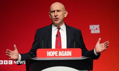 Johm Healey has his arms out in a gesture while speaking at a podium in front of a red backdrop.