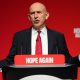Johm Healey has his arms out in a gesture while speaking at a podium in front of a red backdrop.
