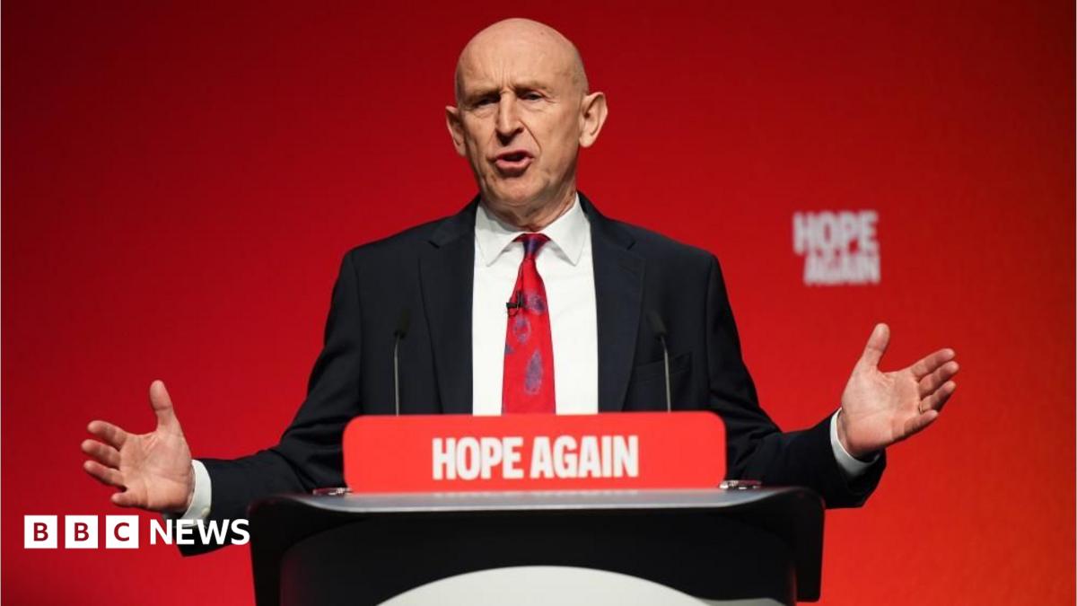 Johm Healey has his arms out in a gesture while speaking at a podium in front of a red backdrop.