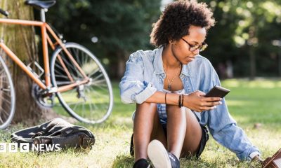 Student sits on the grass with a phone in her hand, a tablet computer on the grass and a bike resting on a tree behind her.