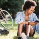 Student sits on the grass with a phone in her hand, a tablet computer on the grass and a bike resting on a tree behind her.