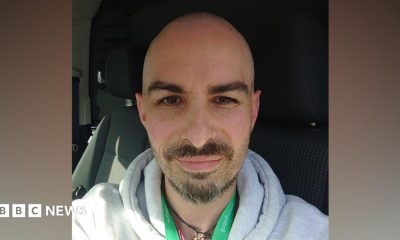 Man with short hair, beard and nose ring. He is wearing a grey hoody with a green lanyard. He is seated in a vehicle.