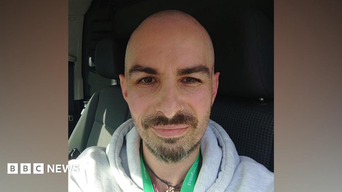 Man with short hair, beard and nose ring. He is wearing a grey hoody with a green lanyard. He is seated in a vehicle.