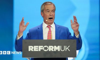 Nigel Farage holds up his hands as he gives his conference speech from behind a podium with Reform UK's logo.