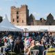 Crowds of people, some of them seated, with marquees in the distance and a castle behind them