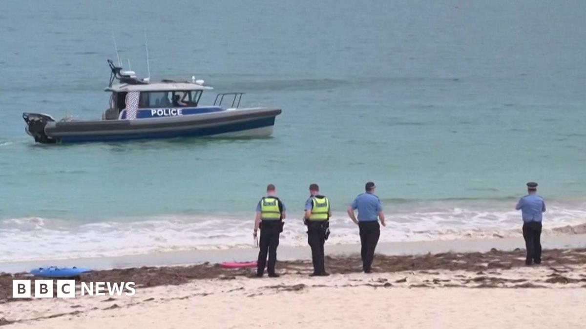 Police officers stand on Sorrento Beach while a police boat patrols offshore during search and recovery operations after a fatal shark attack