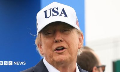 U.S. President Donald Trump attends the Amgen Irish Open at Trump International Golf Links in Doonbeg, Ireland. He is wearing a white USA baseball cap, navy blazer with a white shirt. He has his arms outstretched.