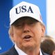 U.S. President Donald Trump attends the Amgen Irish Open at Trump International Golf Links in Doonbeg, Ireland. He is wearing a white USA baseball cap, navy blazer with a white shirt. He has his arms outstretched.