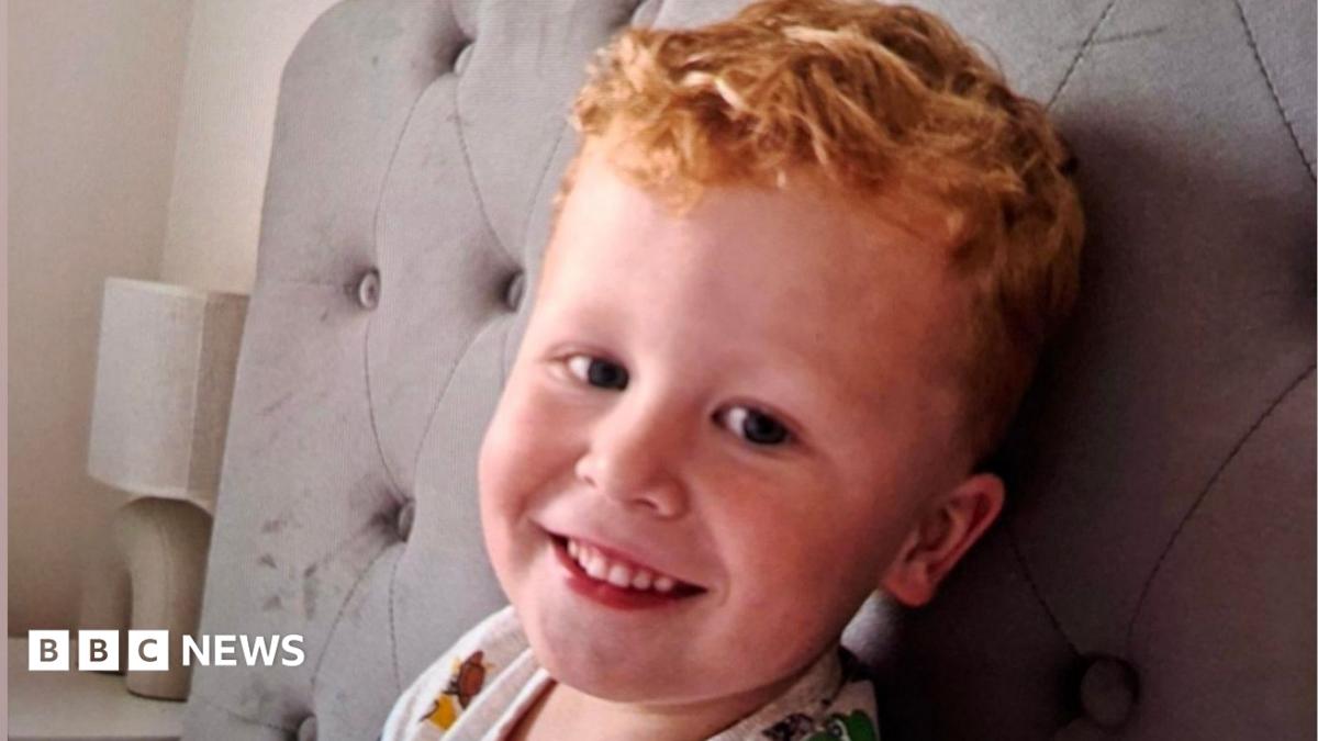 Three-year-old Noah Woods, with light hair and wearing a brightly coloured top, smiles at the camera.