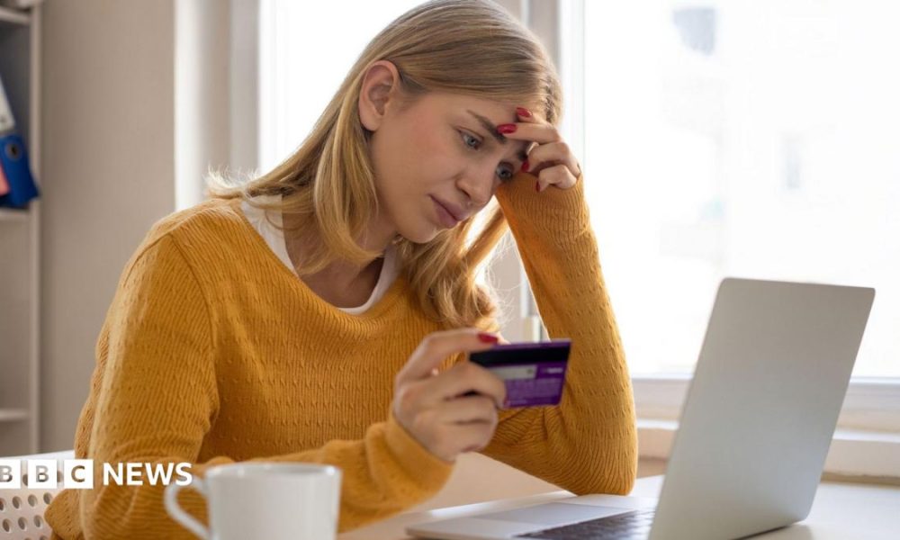 A woman sits at her laptop while holding her bank card in her right hand. She looks deep in thought, with her left hand on the side of her head. She has blonde hair and is wearing a yellow jumper.