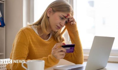 A woman sits at her laptop while holding her bank card in her right hand. She looks deep in thought, with her left hand on the side of her head. She has blonde hair and is wearing a yellow jumper.