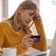 A woman sits at her laptop while holding her bank card in her right hand. She looks deep in thought, with her left hand on the side of her head. She has blonde hair and is wearing a yellow jumper.
