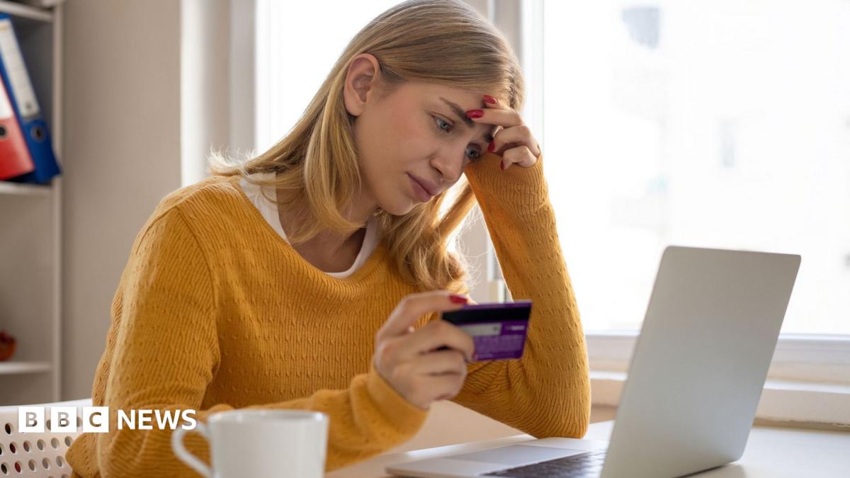 A woman sits at her laptop while holding her bank card in her right hand. She looks deep in thought, with her left hand on the side of her head. She has blonde hair and is wearing a yellow jumper.