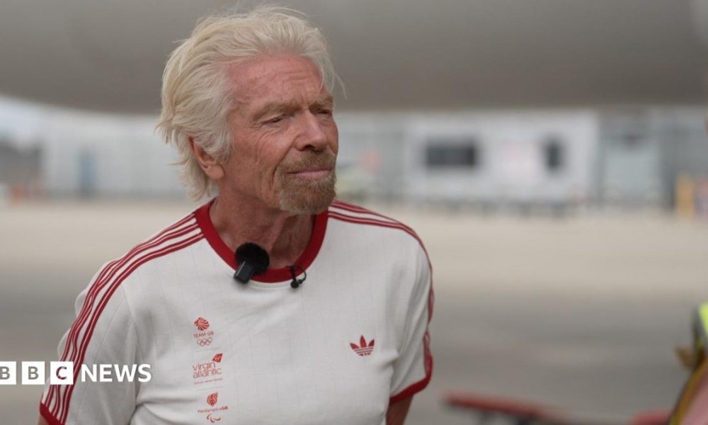 Richard Branson wearing a white t-shirt with the Team GB, Virgin Atlantic, and Paralympics GB logos in the right side of the chest with red lettering, an Adidas logo on the left side of the chest in red, and three red stripes on each arm. He is standing on the runway of Heathrow Airport. Behind him are a Virgin Atlantic plane and the terminal buildings in soft focus.