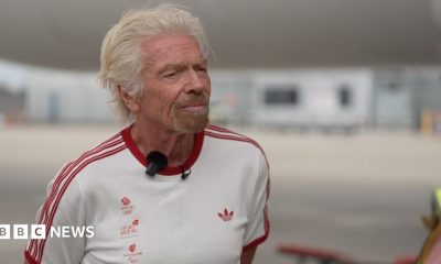 Richard Branson wearing a white t-shirt with the Team GB, Virgin Atlantic, and Paralympics GB logos in the right side of the chest with red lettering, an Adidas logo on the left side of the chest in red, and three red stripes on each arm. He is standing on the runway of Heathrow Airport. Behind him are a Virgin Atlantic plane and the terminal buildings in soft focus.