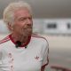 Richard Branson wearing a white t-shirt with the Team GB, Virgin Atlantic, and Paralympics GB logos in the right side of the chest with red lettering, an Adidas logo on the left side of the chest in red, and three red stripes on each arm. He is standing on the runway of Heathrow Airport. Behind him are a Virgin Atlantic plane and the terminal buildings in soft focus.