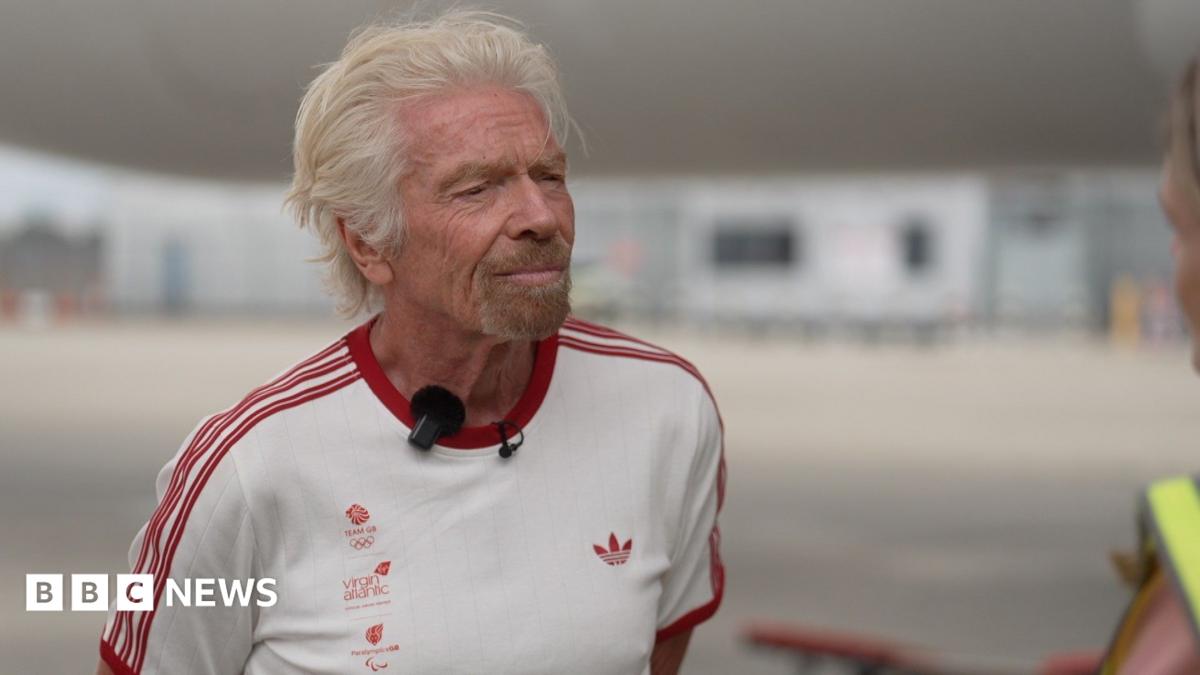 Richard Branson wearing a white t-shirt with the Team GB, Virgin Atlantic, and Paralympics GB logos in the right side of the chest with red lettering, an Adidas logo on the left side of the chest in red, and three red stripes on each arm. He is standing on the runway of Heathrow Airport. Behind him are a Virgin Atlantic plane and the terminal buildings in soft focus.