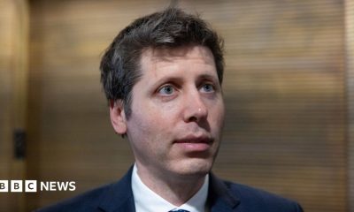 OpenAI CEO Sam Altman standing before a lobby of elevators answering questions from reporters.