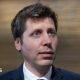 OpenAI CEO Sam Altman standing before a lobby of elevators answering questions from reporters.