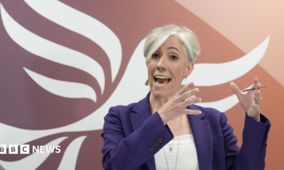 Daisy Cooper speaking on stage in front of a giant orange and white Liberal Democrat bird logo. She is gesticulating with her hands, wearing a purple jacket over a white top, and gold jewellery.