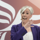 Daisy Cooper speaking on stage in front of a giant orange and white Liberal Democrat bird logo. She is gesticulating with her hands, wearing a purple jacket over a white top, and gold jewellery.