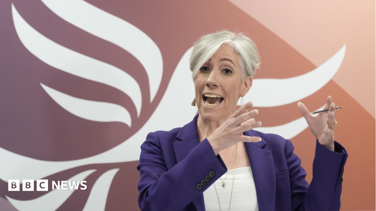 Daisy Cooper speaking on stage in front of a giant orange and white Liberal Democrat bird logo. She is gesticulating with her hands, wearing a purple jacket over a white top, and gold jewellery.