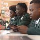 Students in green school uniforms sit at desks reading books during a classroom lesson. Pencil pots and study materials are visible on the tables, with a noticeboard and classroom displays in the background.