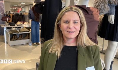 A woman with long blonde hair and wearing a black top and green jacket is standing inside a clothes shop with dressed mannequins and rows of clothes on hangars in the background.