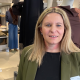 A woman with long blonde hair and wearing a black top and green jacket is standing inside a clothes shop with dressed mannequins and rows of clothes on hangars in the background.