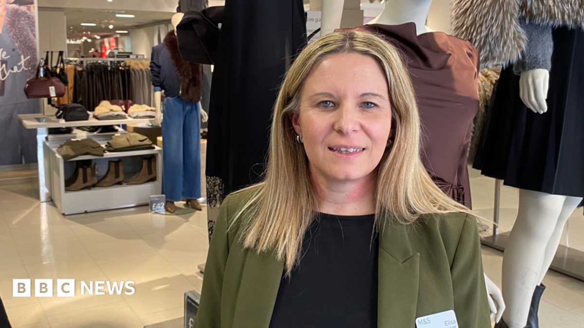 A woman with long blonde hair and wearing a black top and green jacket is standing inside a clothes shop with dressed mannequins and rows of clothes on hangars in the background.