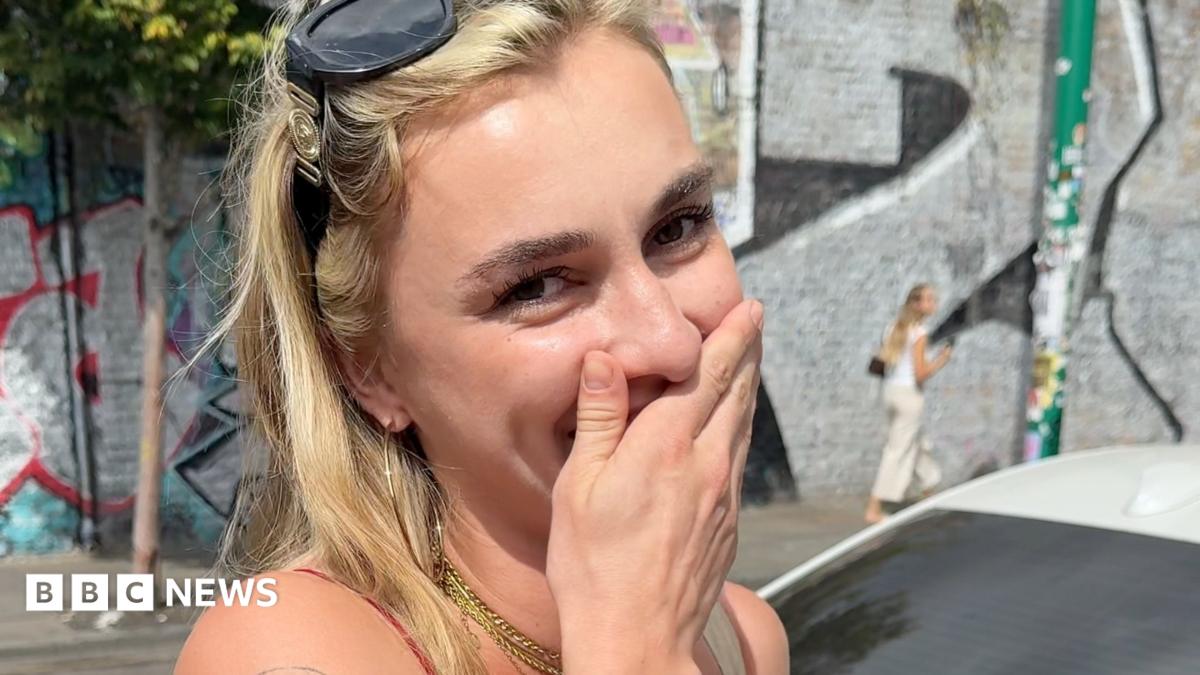 A woman holds her hand over her mouth in the street. She has blonde hair and sunglasses on her head