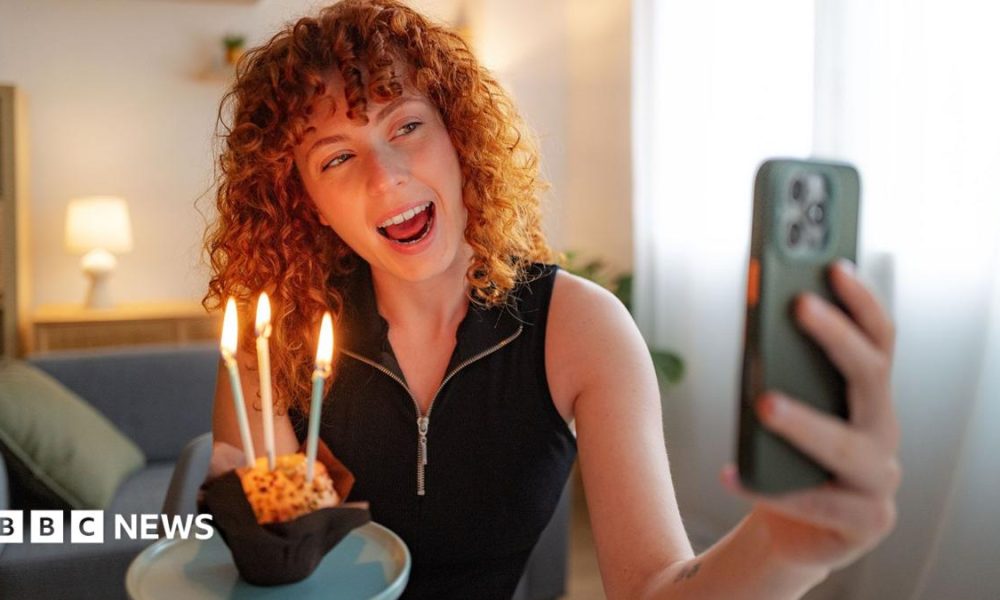A young woman holding a pink birthday cake with five candles on, wearing a party hat and striped shirt, celebrating her birthday, with balloons seen hanging behind her
