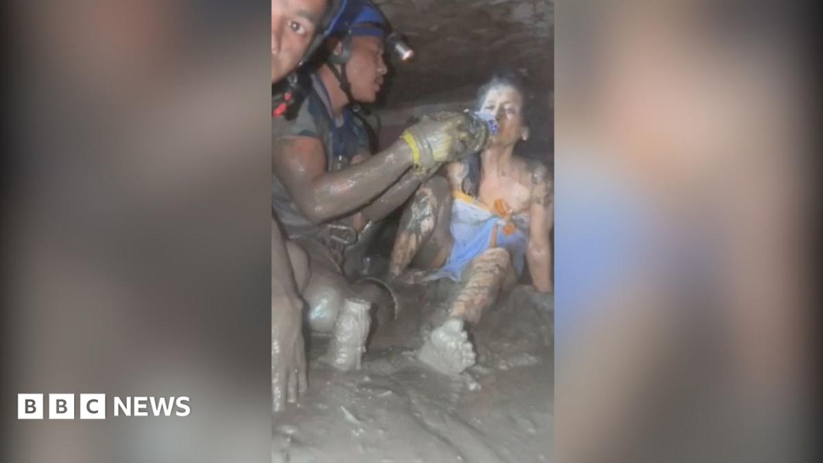 A woman lying in mud being fed water from a bottle by rescuers.