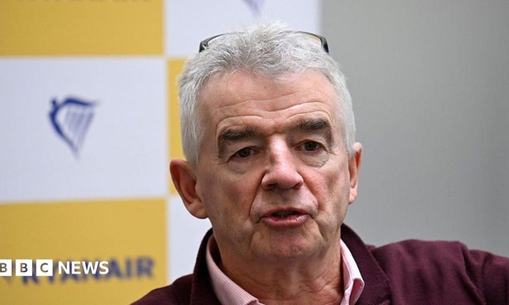 A close-up Michael O'Leary, chief executive of Ryanair, wearing a pink and white chequered shirt and burgundy jumper. Behind him, the Ryanair logo is on a printed background in soft focus.
