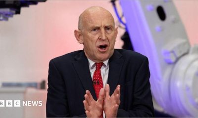 Chancellor John Healey speaking in Coventry in front of purple and grey coloured equipment. He is wearing a dark suit, white short and red tie