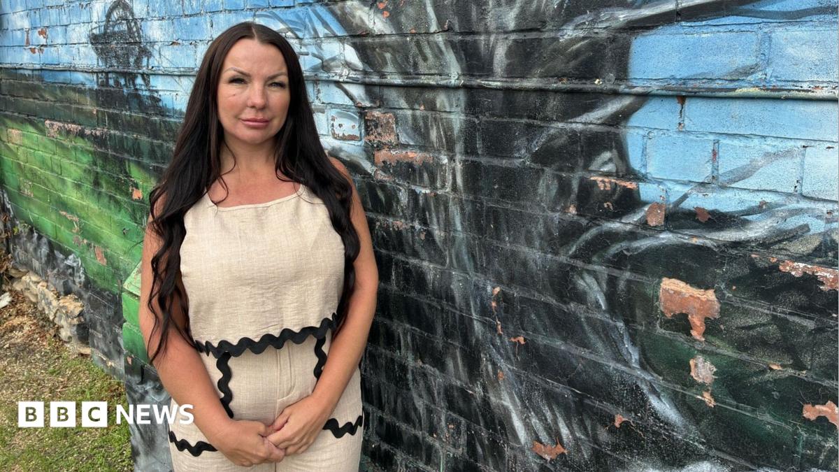 A woman with long black hair is wearing a beige sleeveless top and beige trousers with a black ribbon trim. She is standing in front of a brick wall which has a tree mural painted on it