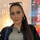 A woman with long, dark hair that is tied up in a ponytail. She is wearing a grey T-shirt and black jacket. She is looking into the camera with a straight face. She is wearing large, gold hoop earring and has a pair of sunglasses on top of her head.