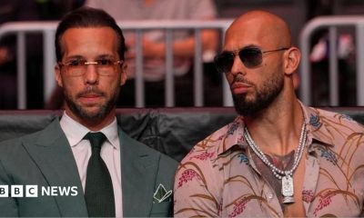 Tristan Tate is wearing sunglasses and is dressed in a green suit and tie and white shirt. Andrew is to his right in sunglasses and an open necked shirt.