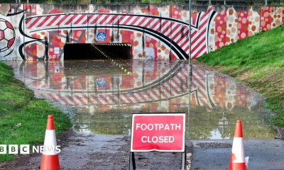 A flooded footpath leading to an underpath with warning cones and a "footpath closed" sign