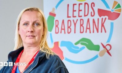A woman with shoulder-length blonde hair and blue eyes looks into the camera with a neutral expression. She is wearing a dark blue fleece jacket and has a red lanyard around her neck. In the background is a large sign with Leeds Baby Bank on it, along with images of hands, dummies, prams and baby bottles
