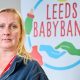 A woman with shoulder-length blonde hair and blue eyes looks into the camera with a neutral expression. She is wearing a dark blue fleece jacket and has a red lanyard around her neck. In the background is a large sign with Leeds Baby Bank on it, along with images of hands, dummies, prams and baby bottles