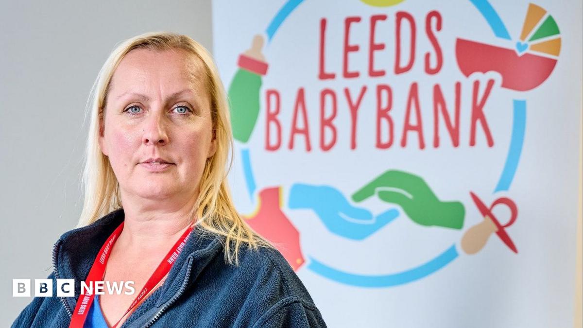 A woman with shoulder-length blonde hair and blue eyes looks into the camera with a neutral expression. She is wearing a dark blue fleece jacket and has a red lanyard around her neck. In the background is a large sign with Leeds Baby Bank on it, along with images of hands, dummies, prams and baby bottles
