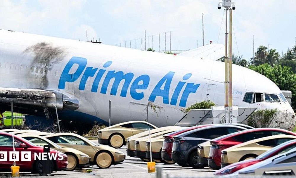 A parking lot filled with Tesla Cybercabs is seen in the foreground as the nose of a Prime Air cargo plane rests on the ground after the aircraft overran the runway and struck cars the previous day at Miami International Airport in Miami, Florida, on September 7, 2026.