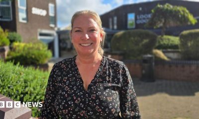 Sally Hellwich has blonde hair tied back and is smiling. She's wearing a black blouse with a colourful floral pattern. She is standing outside in a pedestrianised street with bushes and trees around brown buildings, on a sunny and cloudy day.