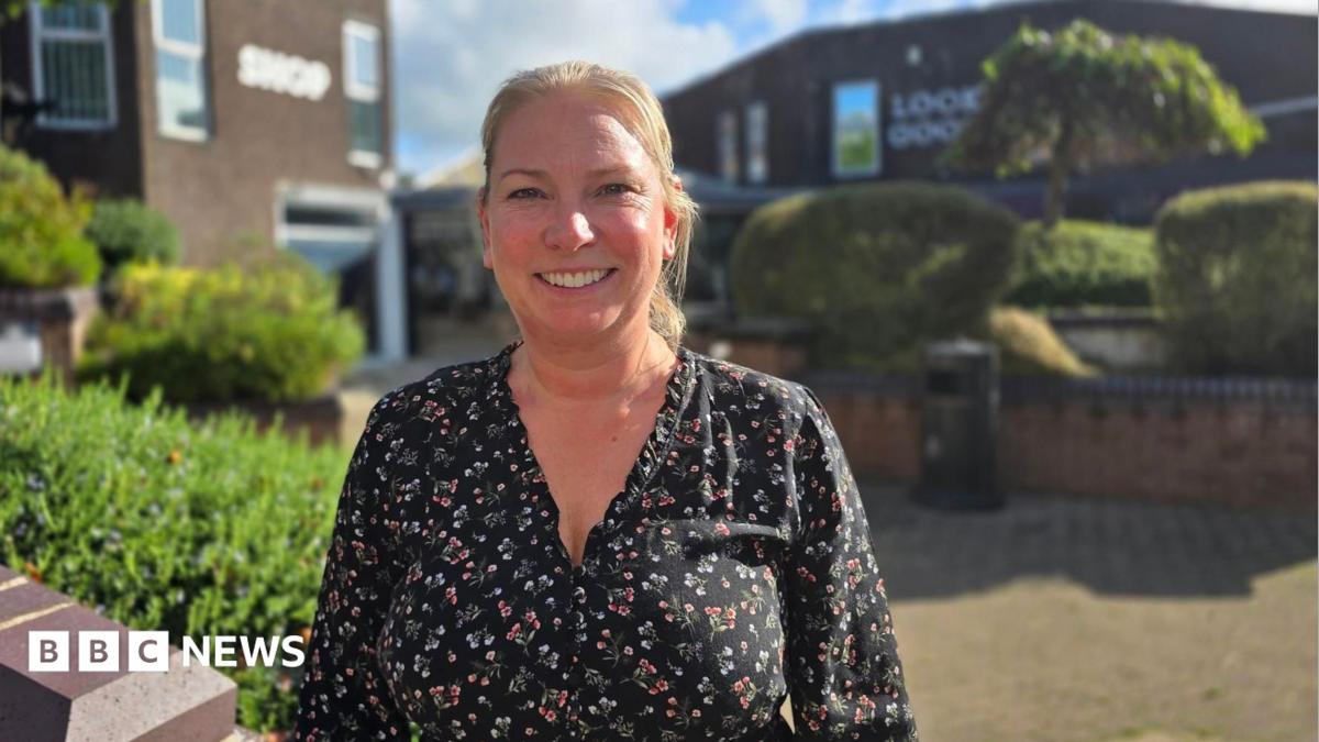 Sally Hellwich has blonde hair tied back and is smiling. She's wearing a black blouse with a colourful floral pattern. She is standing outside in a pedestrianised street with bushes and trees around brown buildings, on a sunny and cloudy day.